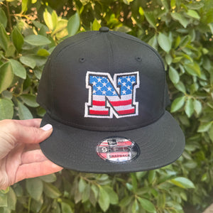 Black cap with American flag logo held in front of green foliage