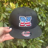 Black cap with American flag logo held in front of green foliage