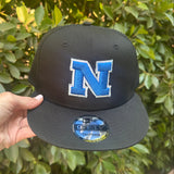 A black New Era 9FIFTY snapback cap with a blue and white embroidered letter 'N' on the front, held by a hand against a background of green foliage.
