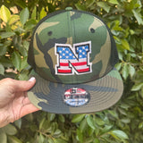 Camouflage cap with American flag logo held in front of green foliage